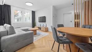 A modern living room with a sofa, a round wooden dining table, and a TV mounted on the wall.