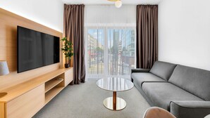 Una camera d'albergo moderna con televisione a schermo piatto, un divano grigio, un tavolino rotondo e un'ampia finestra con tende.
