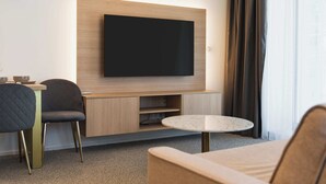 Ein modernes Hotelzimmer mit Flachbildfernseher an einer Holzwand, einem runden Marmortisch, einer Couch und einem Sessel.