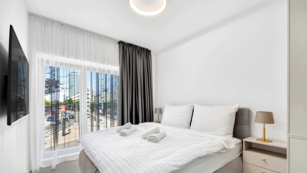 Ein modernes Hotelzimmer mit einem großen Bett, einem an der Wand montierten Fernseher, einem Nachttisch mit Lampe und Blick auf die Stadt durch große Fenster.