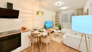 Een moderne woonkamer met een flatscreen televisie, een eethoek met een tafel voor vier personen, een bank en een groot raam met gordijnen.