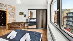 Ein modernes Hotelzimmer mit großem Fenster, einem Bett mit Handtüchern, einem braunen Ledersofa, einem kleinen Tisch und einer Wand mit Zeitungsartikeln.