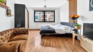 Ein modernes Schlafzimmer mit großem Fenster, einem Bett, einem braunen Ledersofa, einem Fernseher und einer Pflanze auf einem kleinen Tisch.