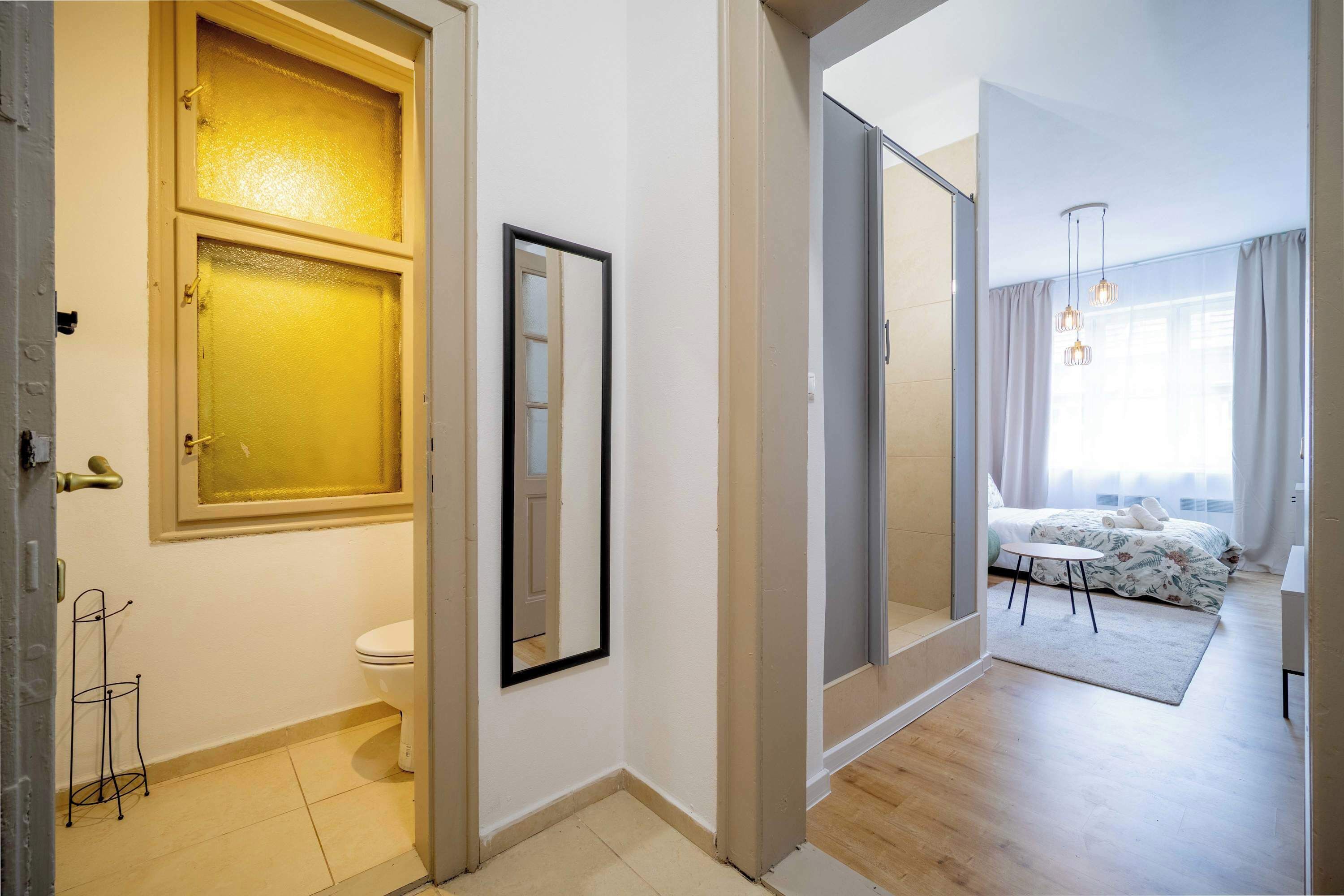 Una habitación de hotel moderna con baño, una cama con cubrecama floral, una mesita pequeña y un ventanal con cortinas.