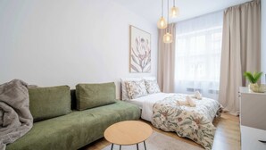 A modern bedroom with a green sofa, a bed with floral bedding, a small round table, and a ceiling light fixture. - Elegant Studio in Historical Centrum | AC & Netflix (Košice I)