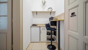 A compact kitchen with a white refrigerator, a small sink, and a wooden countertop. There is a black chair and a shelf with a clock and decorative items. - Elegant Studio in Historical Centrum | AC & Netflix (Košice I)