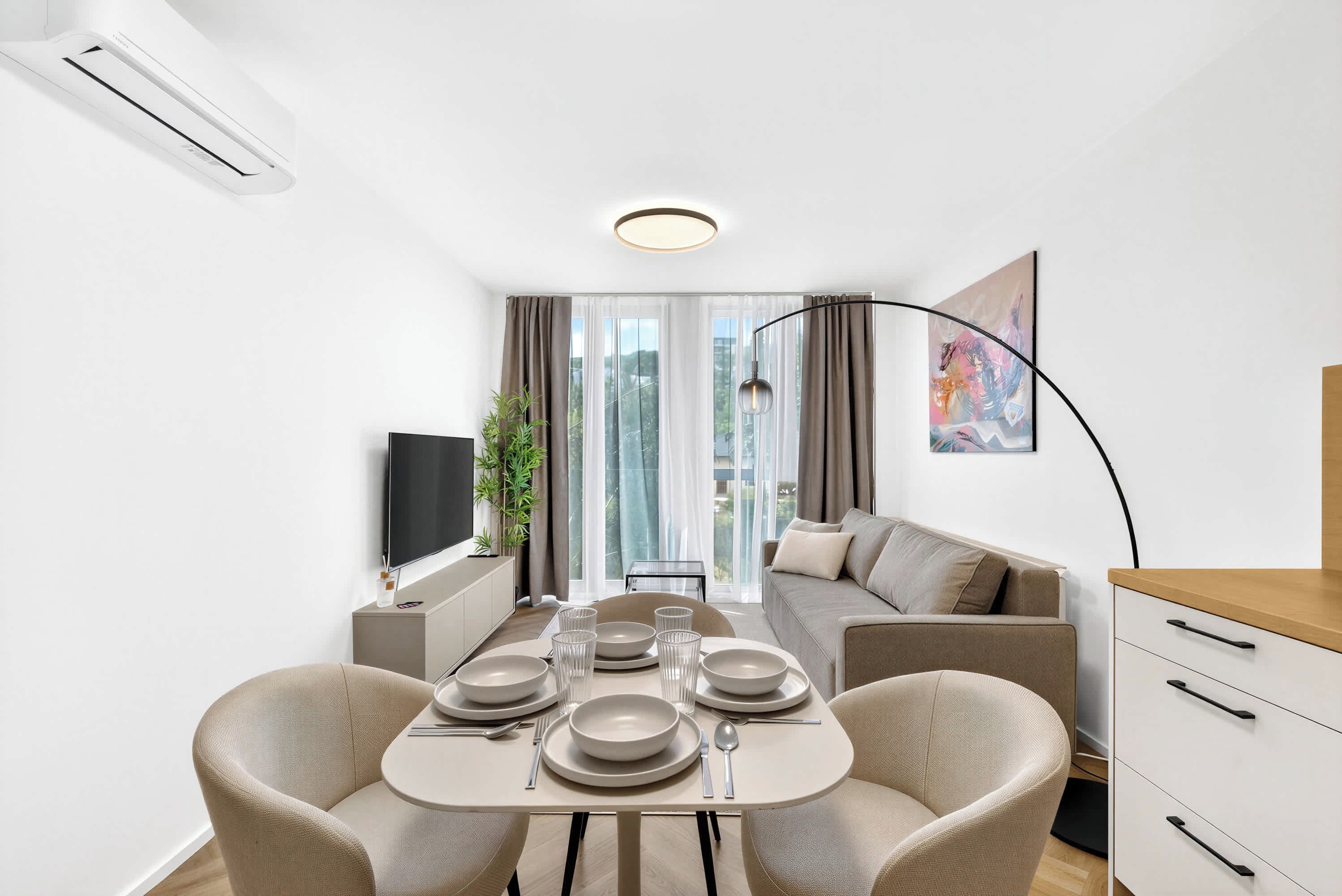 A modern living room with a dining table set for four, a sofa, a TV, and a large window with curtains.