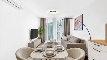 A modern living room with a dining table set for four, a sofa, a TV, and a large window with curtains.
