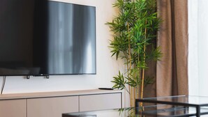 Ein modernes Wohnzimmer mit einem wandmontierten Flachbildfernseher, einem Glastisch, einer Bambuspflanze und einem braunen Vorhang.