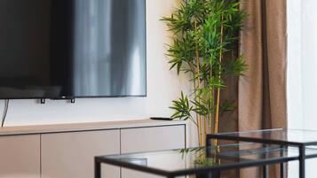 A modern living room with a flat-screen TV mounted on the wall, a glass coffee table, a bamboo plant, and a brown curtain.