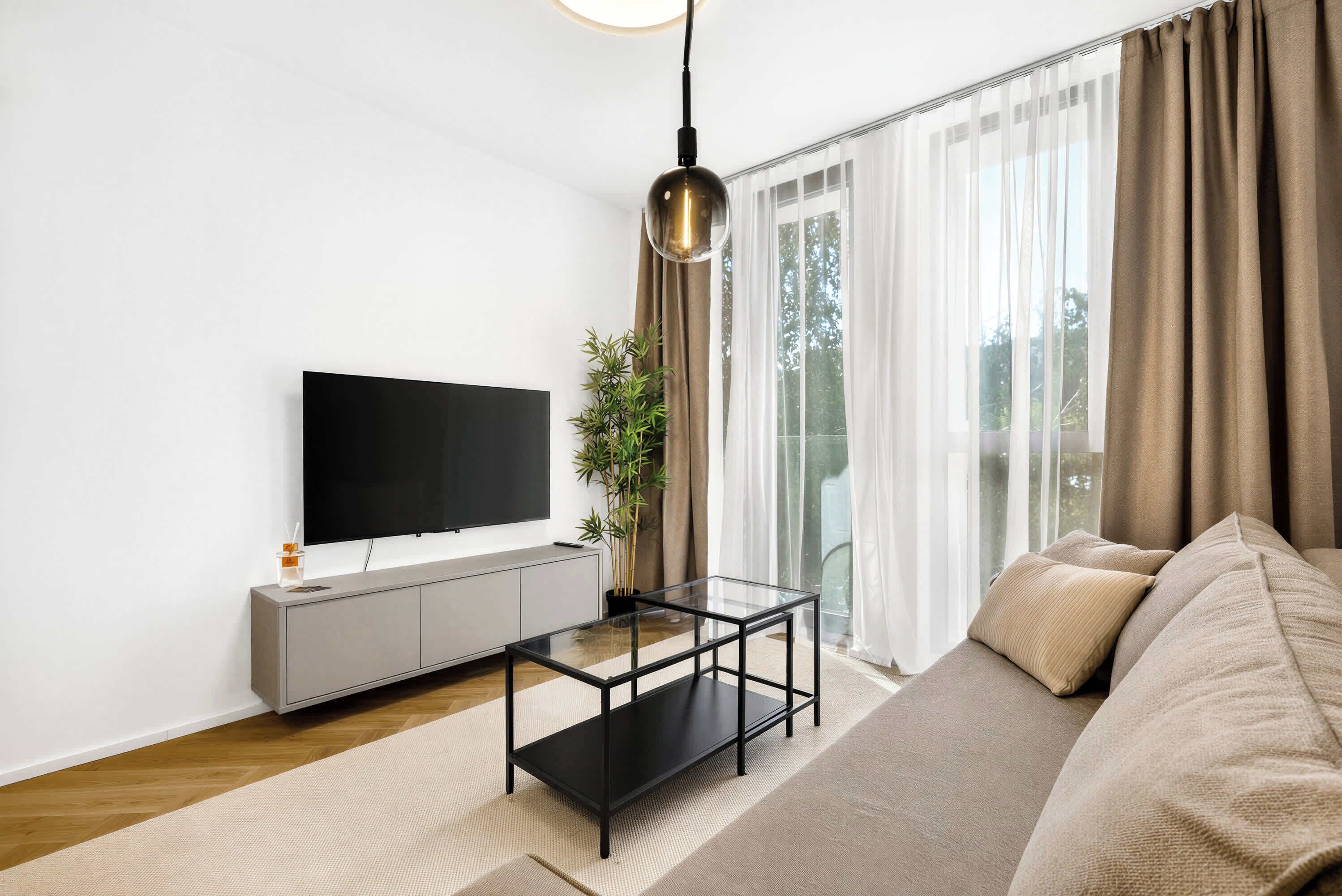 Una sala de estar moderna con un televisor de pantalla plana, una mesa de centro de vidrio, un sofá y un ventanal con cortinas.