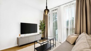 Ein modernes Wohnzimmer mit Flachbildfernseher, Glastisch, Sofa und einem großen Fenster mit Vorhängen.