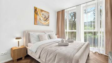 A bedroom with a bed, a bedside table, a lamp, a painting, and a window with curtains.
