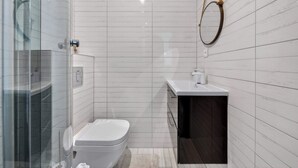 Et moderne badeværelse med en glasbrusekabine, et hvidt toilet og en mørk kommode med spejl.