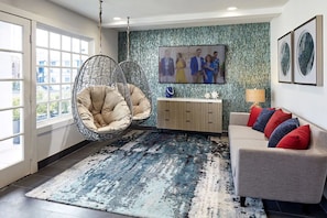 Un salon moderne avec un fauteuil suspendu, un canapé agrémenté de coussins colorés, un téléviseur fixé au mur et un tapis à motifs.