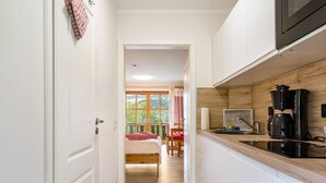 Un pasillo estrecho que conduce a un dormitorio con cama, una mesita y una silla. A la derecha se observa una zona de cocina con microondas y cafetera.