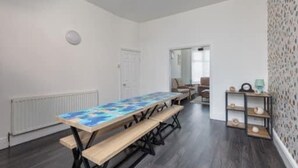 Une pièce avec une longue table en bois, des bancs, un radiateur, une porte et une étagère remplie d’objets.