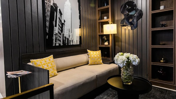 A modern hotel room with a sofa, a round coffee table, a vase with flowers, a wall-mounted lamp, and a large cityscape painting.