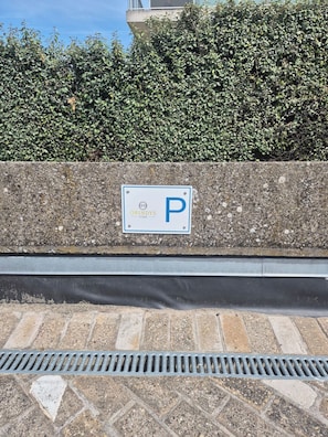 Senyal de pàrquing amb una 'P' a una paret de pedra, una reixa metàl·lica i un arbust al fons.