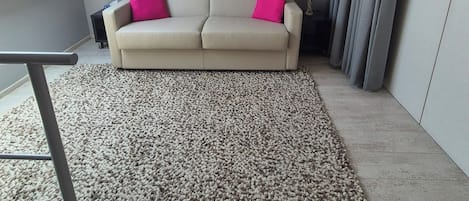 Ein modernes Wohnzimmer mit hoher Decke, einer Couch mit zwei Kissen, einem großen Teppich, einer Stehlampe und einem Gemälde an der Wand.