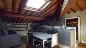 Ein modernes Loft-Apartment mit freiliegenden Holzbalken, einem Essbereich mit grauem Tisch und Stühlen, einer Küche mit integrierter Kochinsel und einem Oberlicht.