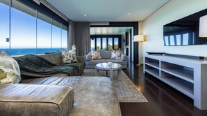 Ein modernes Wohnzimmer mit Sofa, Sessel und Couchtisch, einem Fernseher an der Wand und Blick aufs Meer.