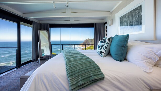 Ein großes Bett mit einer grünen Decke, weißen Kissen und Blick auf das Meer durch große Fenster.