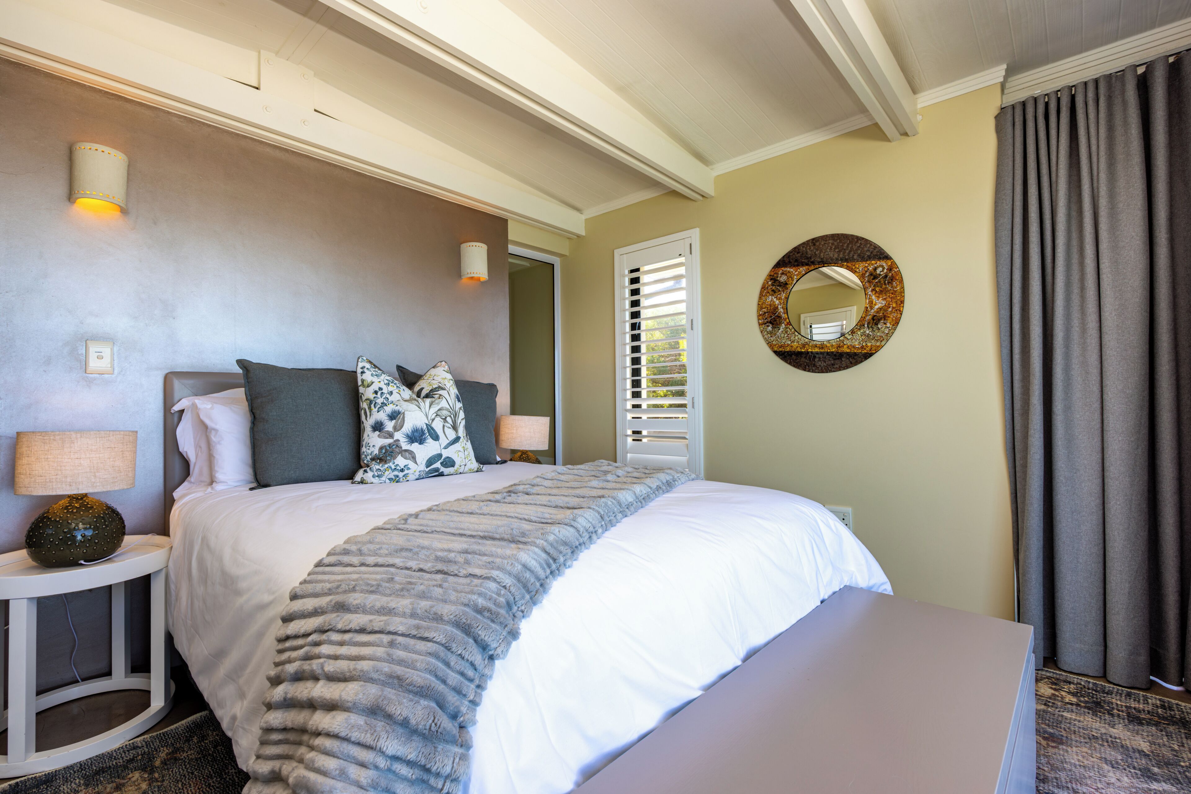A hotel room with a bed, bedside tables, a lamp, a mirror, and a window with blinds.