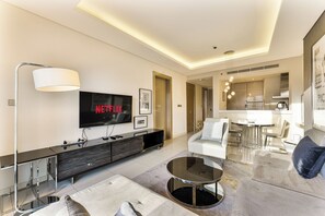 A modern living room with a large flat-screen TV, a sofa, a coffee table, and a dining area with a kitchen in the background. - Amazing 1BR, 10 mins to Burj Khalifa (Dubai)