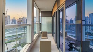Un balcón moderno con amplios ventanales, barandilla de cristal y vistas a un paisaje urbano con edificios y grúas de construcción.