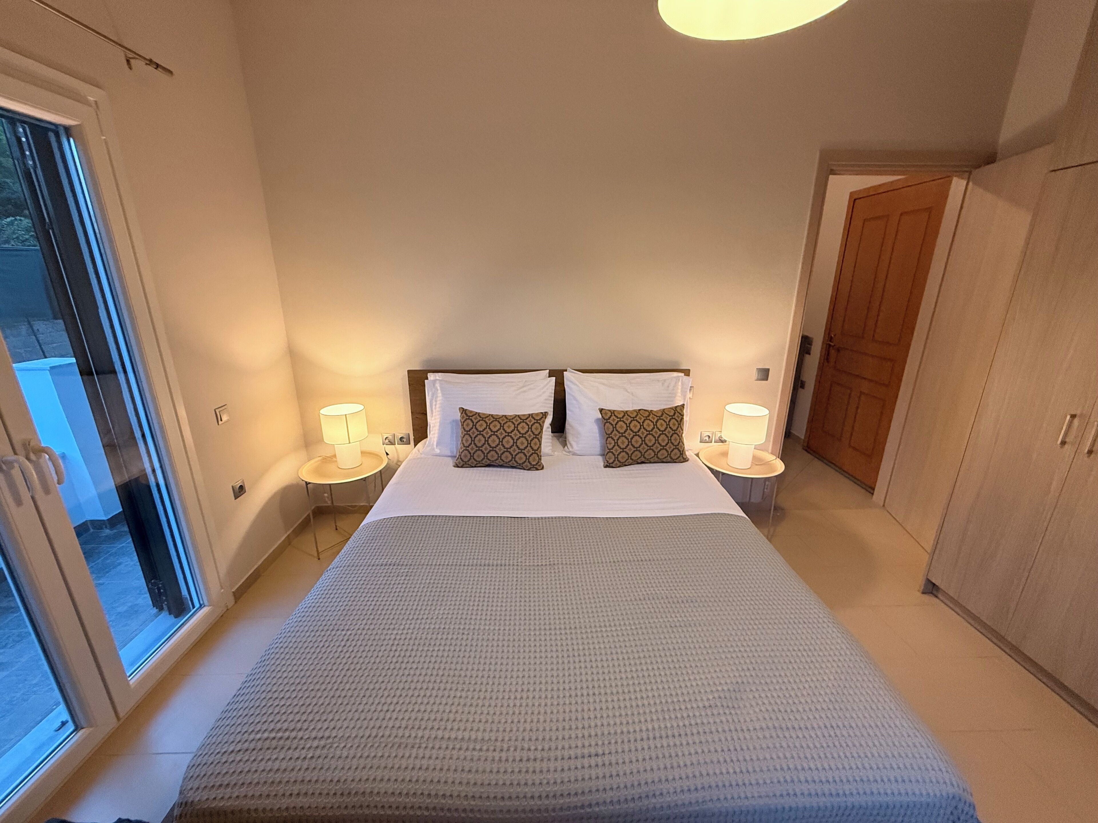 A hotel room with a large bed, two bedside tables with lamps, a wardrobe, and a sliding glass door leading to a balcony.