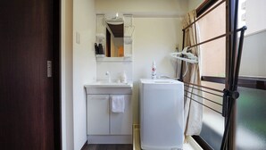 A compact bathroom with a sink, mirror, towel, and a small white appliance, likely a water heater or heater, with a window and curtains. - Guest House WASSO (京都市伏見区深草下川原町)