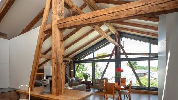 A spacious loft with exposed wooden beams, a large window offering a view of greenery, a coffee machine, and a dining area.