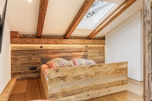 Ein Schlafzimmer im Loft-Stil mit schrägem Dach, Holzbalken, einem Oberlicht und einem Bett mit gemusterten Betttextilien.