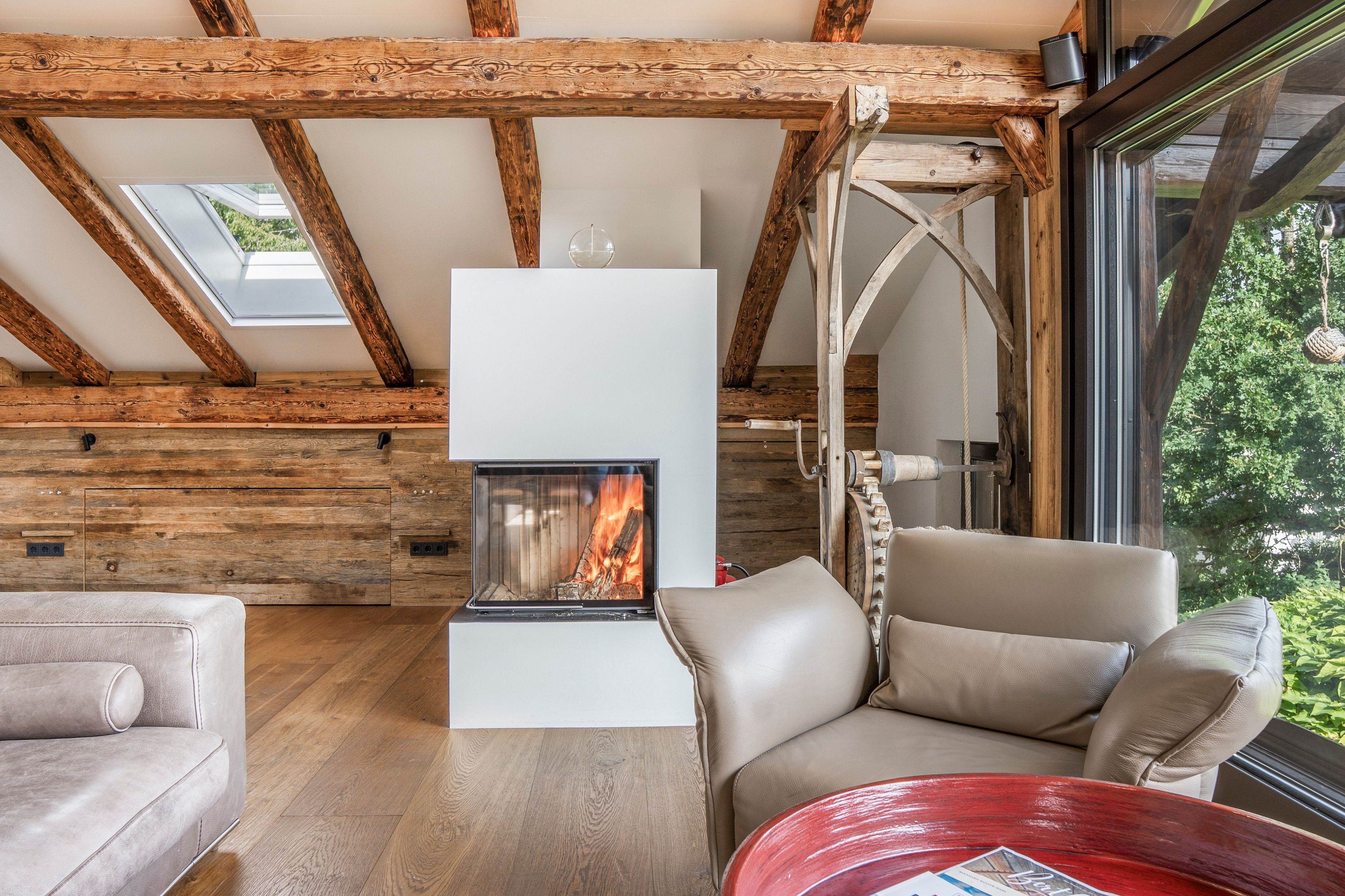 Ein gemütliches Zimmer mit Kamin, Holzbalken, einer Couch und Blick auf Grünflächen.