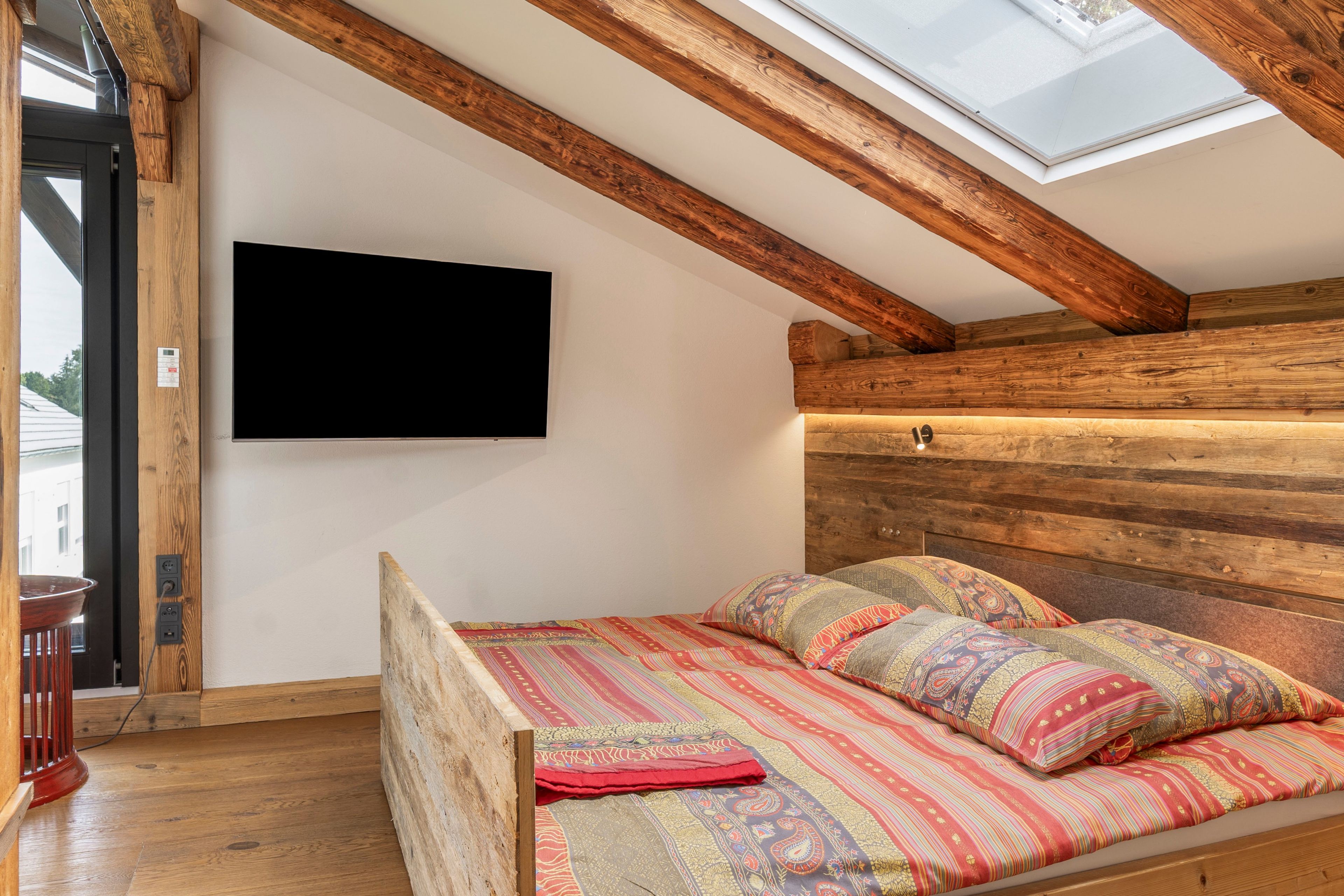 Un dormitorio con techo inclinado, vigas de madera, una cama con ropa de cama estampada y un televisor de pantalla plana montado en la pared.