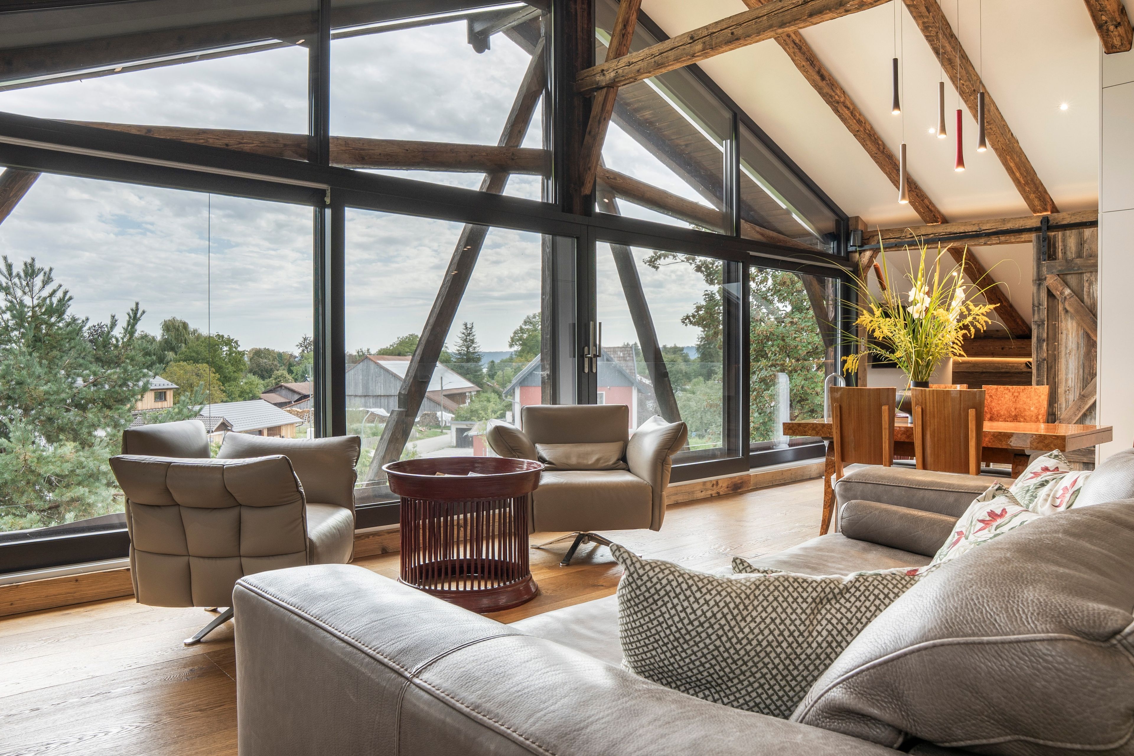 Een ruime woonkamer met grote ramen, een hoog plafond met zichtbare balken en uitzicht op het omliggende landschap.