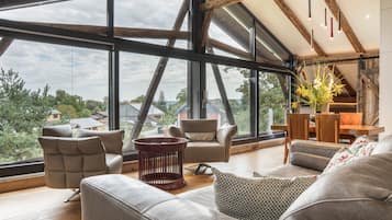 Een ruime woonkamer met grote ramen, een hoog plafond met zichtbare balken en uitzicht op het omliggende landschap.