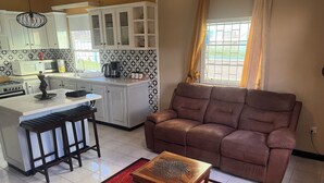 A kitchen with white cabinets, a microwave, a stove, and a coffee maker. There's a brown leather sofa and a wooden table with a patterned rug.
