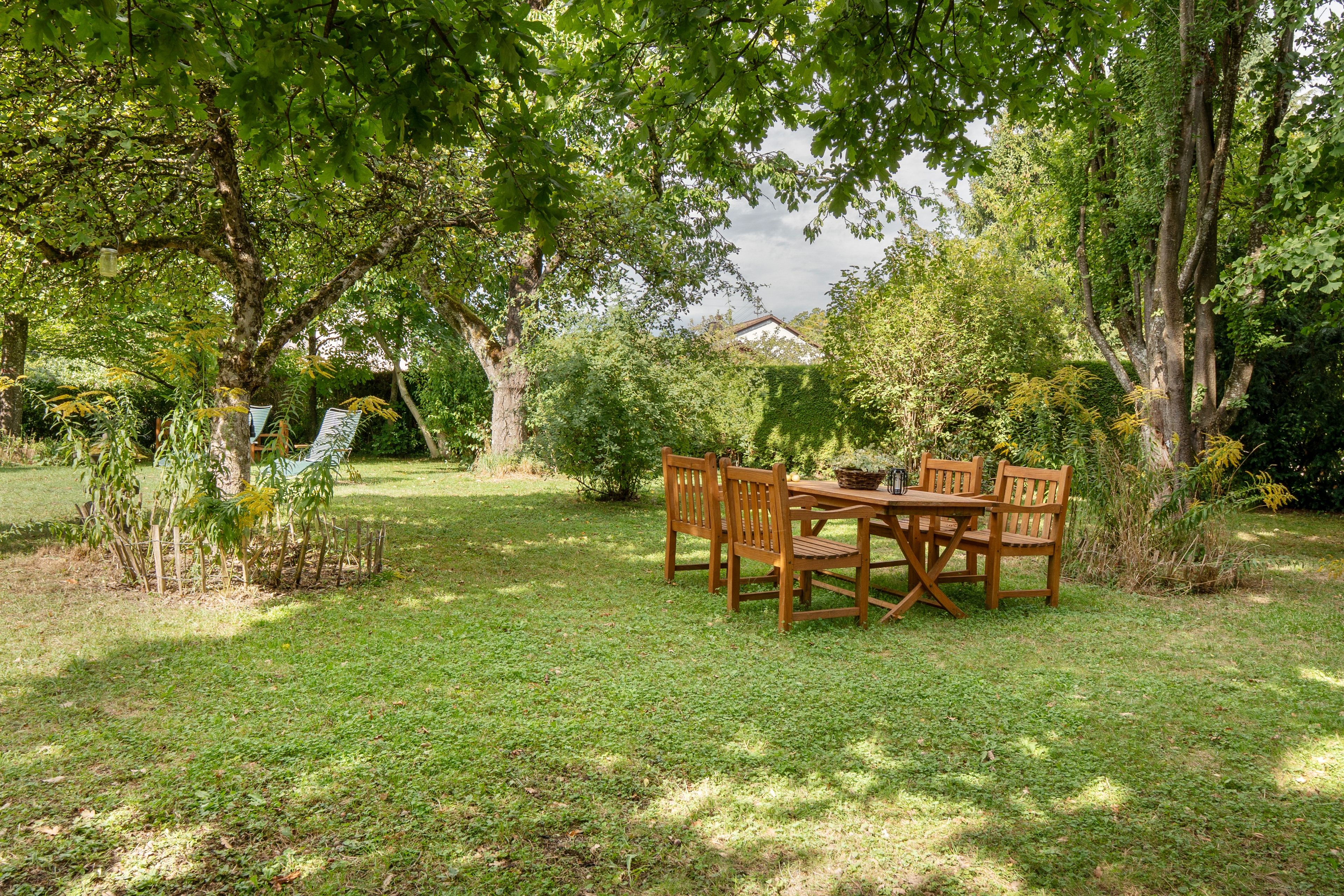 Ein Holztisch mit Stühlen steht in einem grünen Garten mit Bäumen und Blick auf ein Gebäude im Hintergrund.