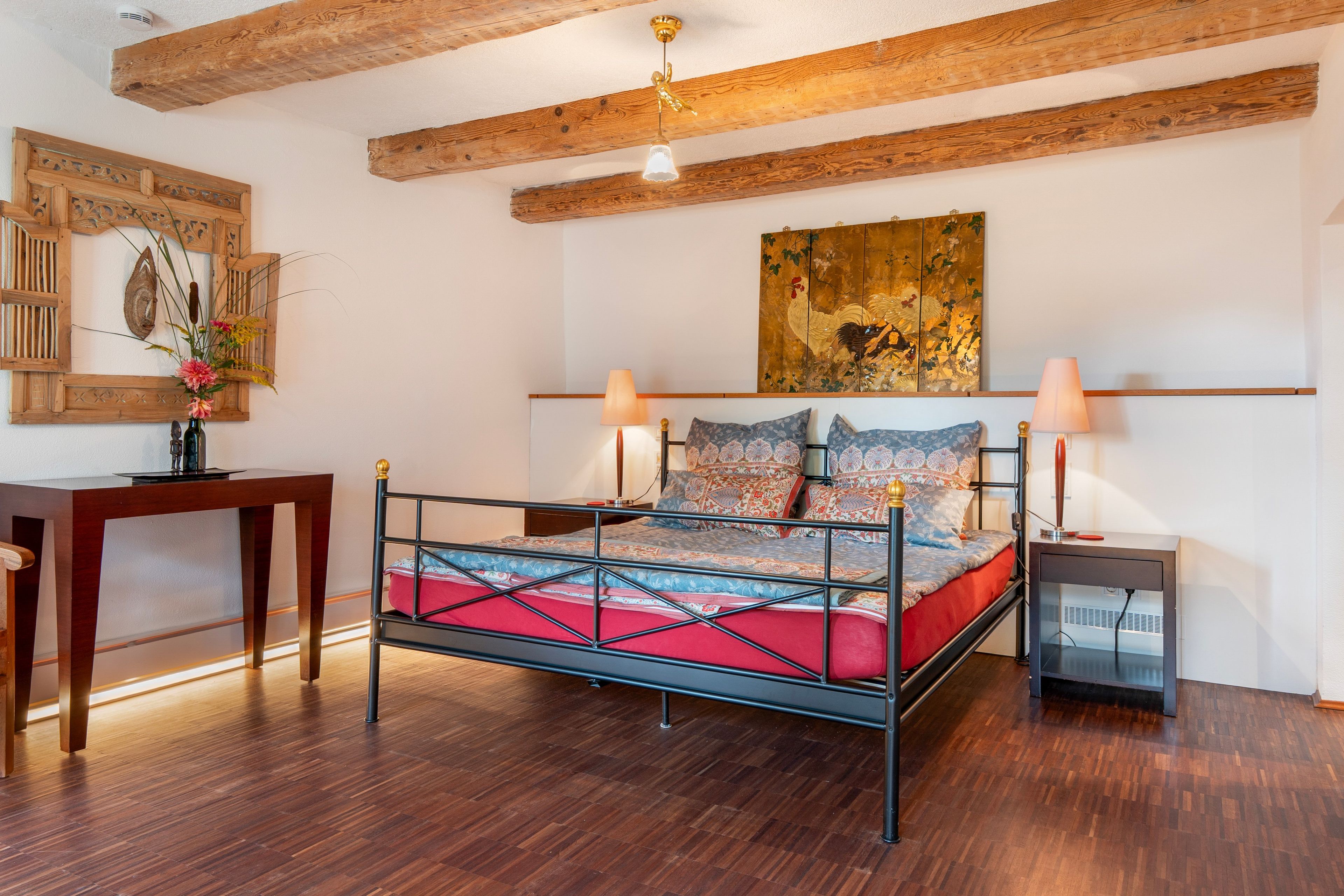 Ein Schlafzimmer mit einem Metallbett, Holzbalken an der Decke, einem Schreibtisch und einem an der Wand befestigten Kunstwerk.