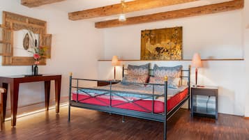 Quarto com cama de estrutura metálica, vigas de madeira no teto, escrivaninha e quadro pendurado na parede.