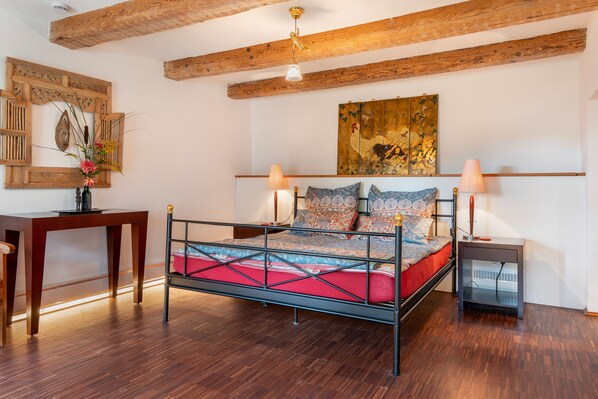A bedroom with a metal-framed bed, wooden beams on the ceiling, a desk, and a wall-mounted artwork.