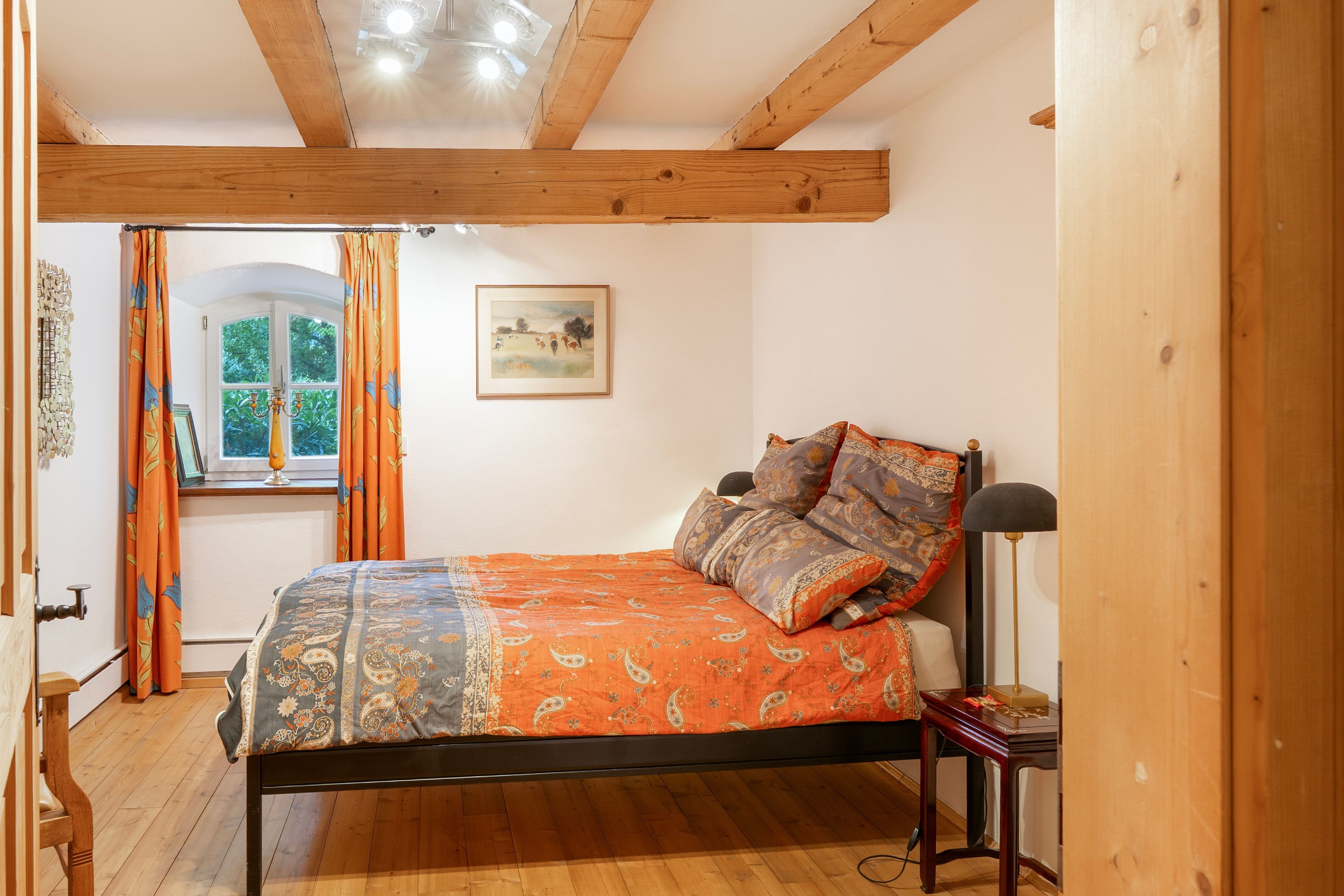 Ein Schlafzimmer mit Holzboden, einem Bett mit gemusterten Betttextilien, einem Fenster mit Vorhängen und einem Bild an der Wand.
