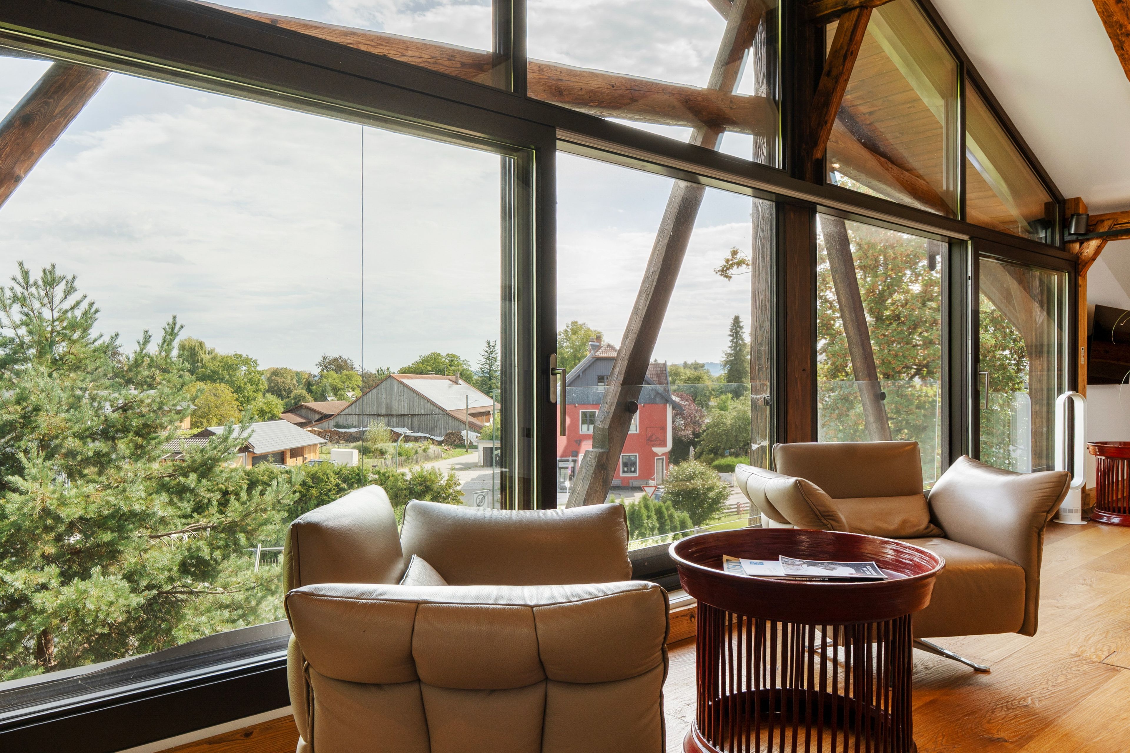 Ein geräumiges Zimmer mit großen Fenstern und Blick auf eine Wohngegend. Zwei Ledersessel und ein Holztisch sind ebenfalls vorhanden.