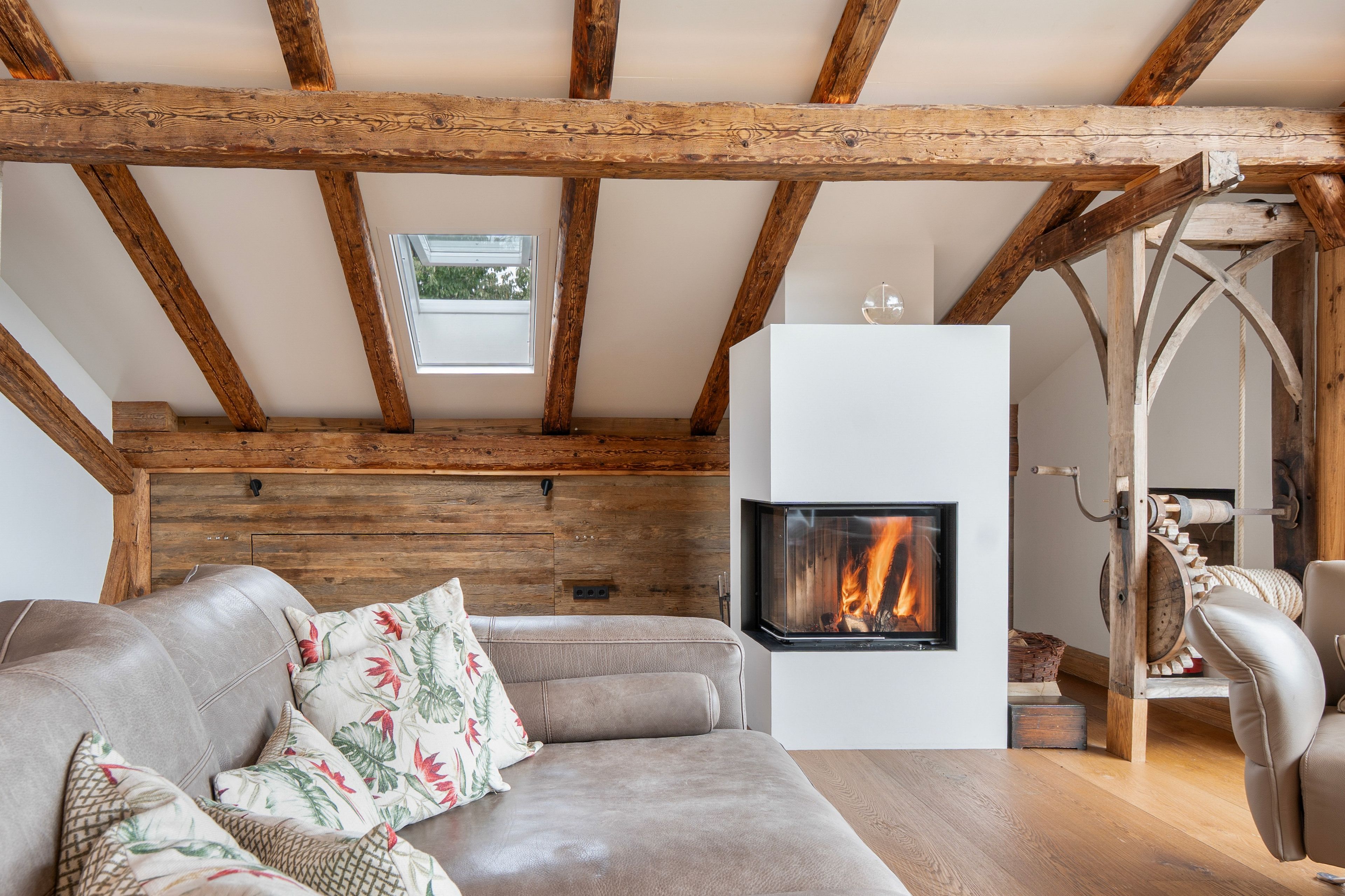 A cozy loft bedroom with exposed wooden beams, a fireplace, a sofa with floral cushions, and a view of the outdoors through a window.