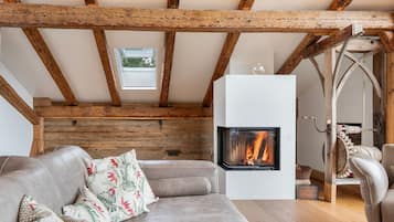 A cozy loft bedroom with exposed wooden beams, a fireplace, a sofa with floral cushions, and a view of the outdoors through a window.