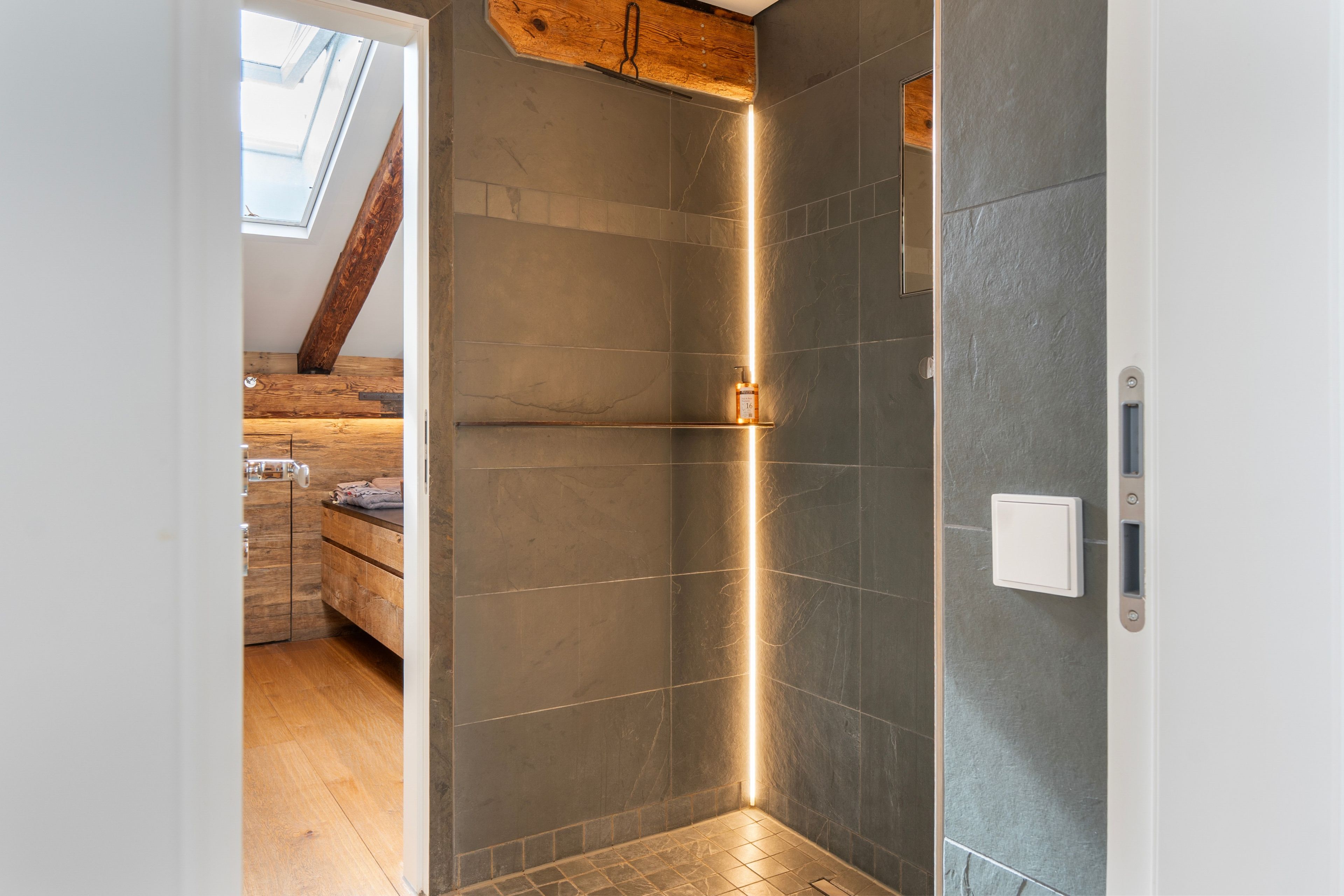 Een moderne badkamer met donkere tegels, een douchegedeelte en een houten plafond.