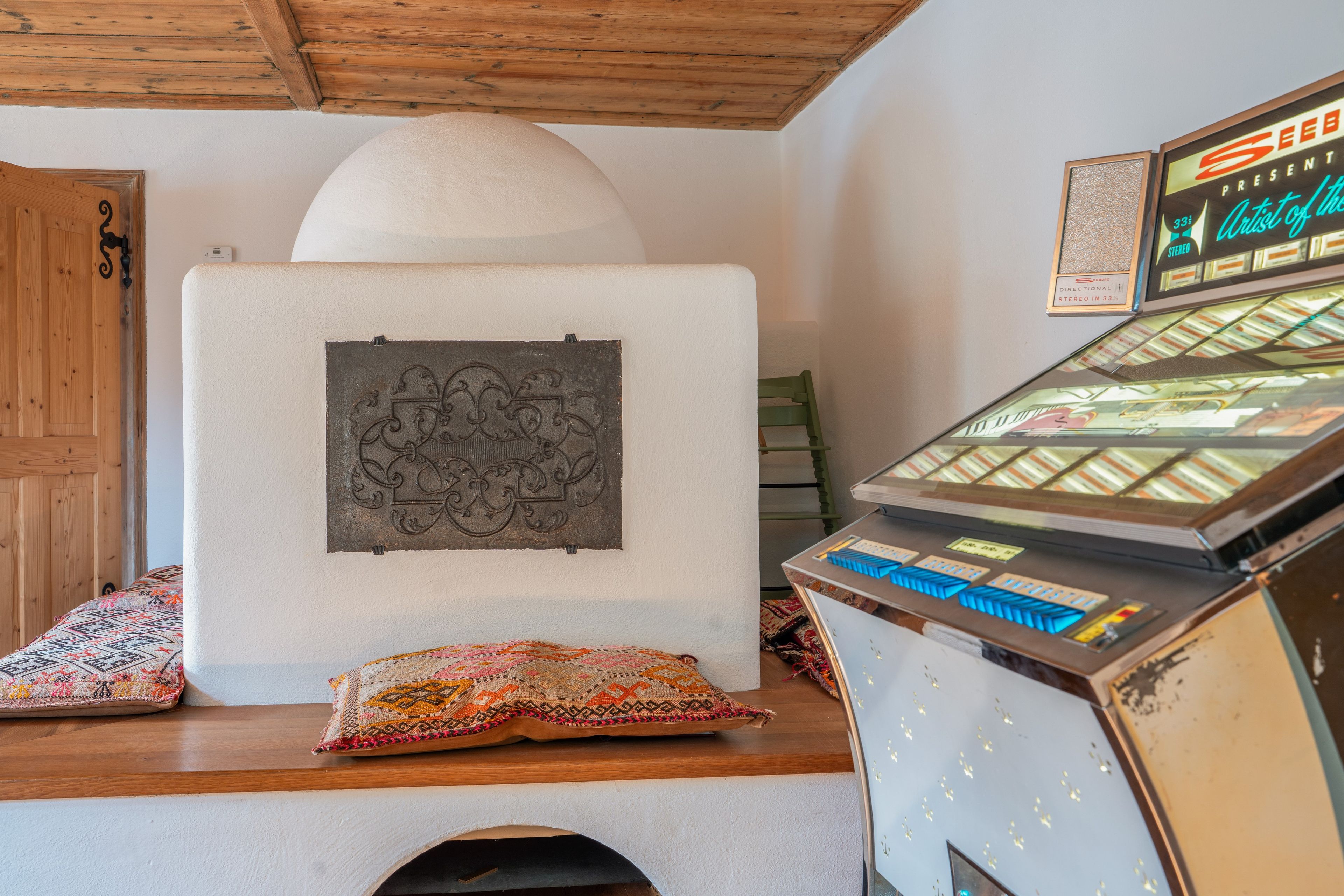 Ein Zimmer mit einer Holztür, einer weißen Wand mit einer verzierten Metallplakette, einem kleinen Tisch mit einem bunten Kissen und einem Vintage-Jukebox.