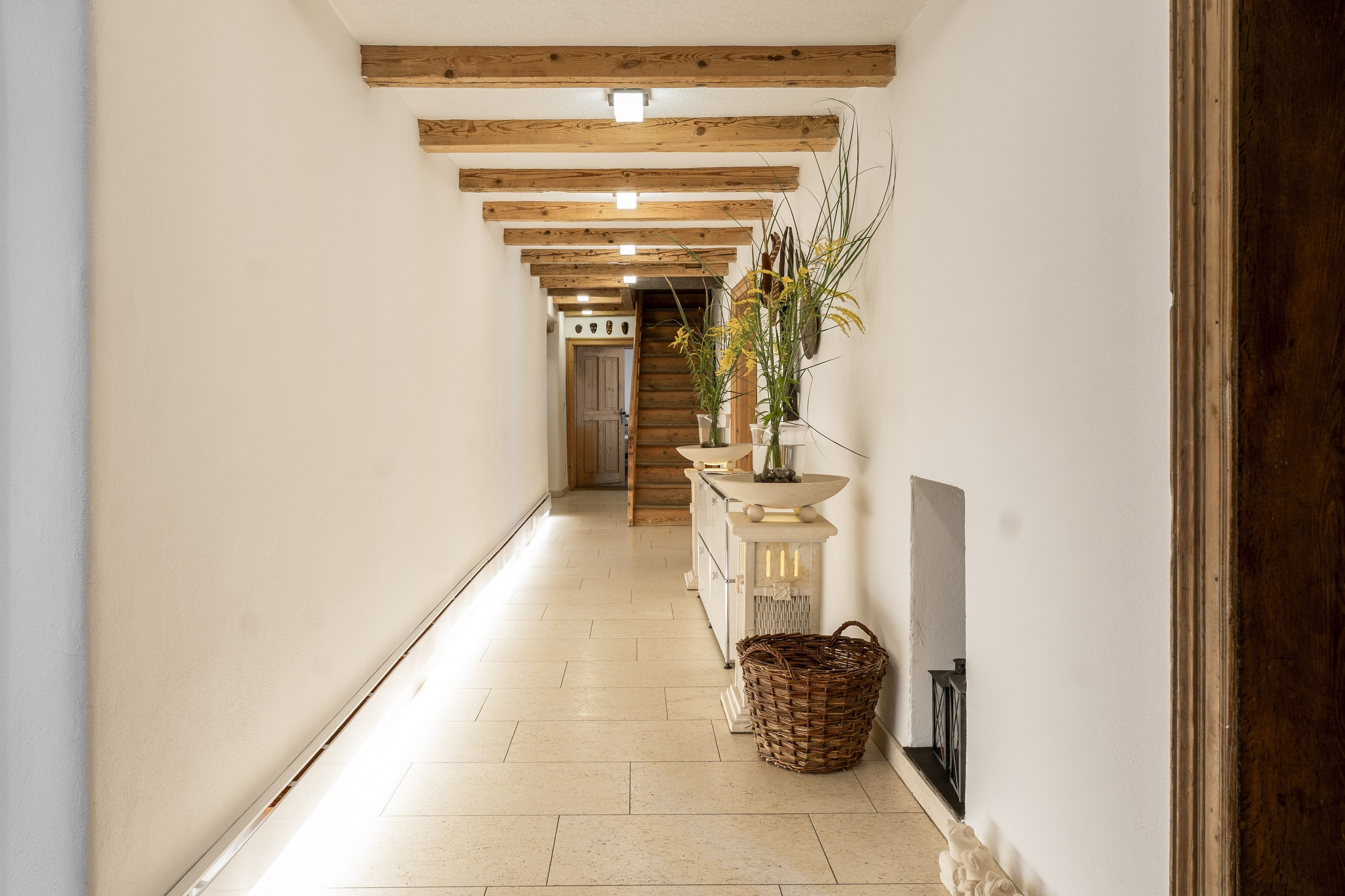A hallway with wooden beams, a white tiled floor, a wicker basket, and a plant in a vase.